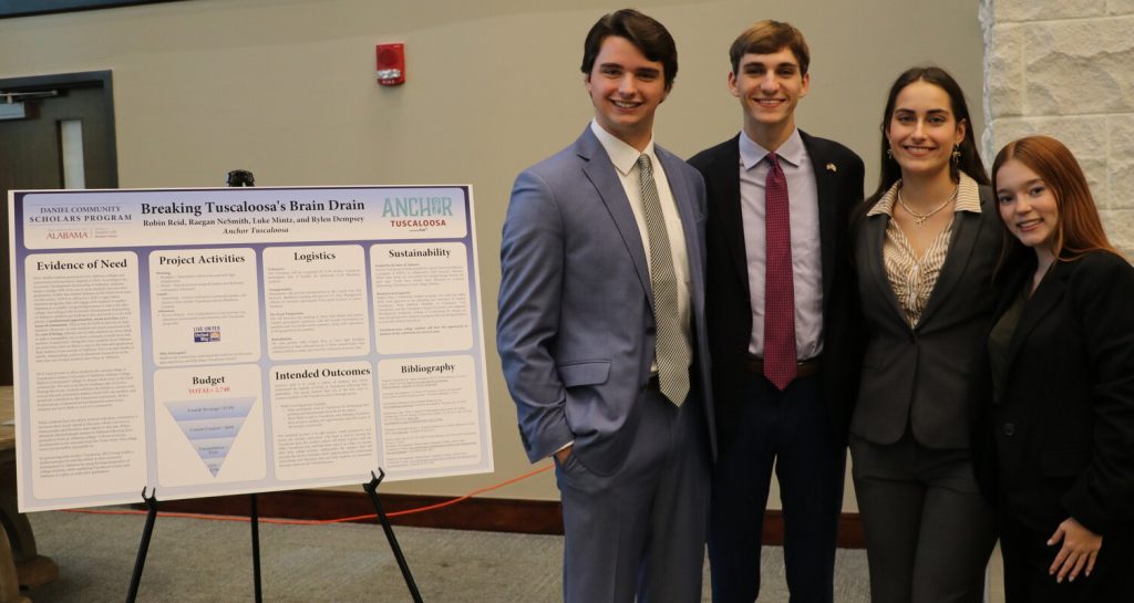 Four Daniel Community Scholars from Team Four stand smiling next to the poster of their project.