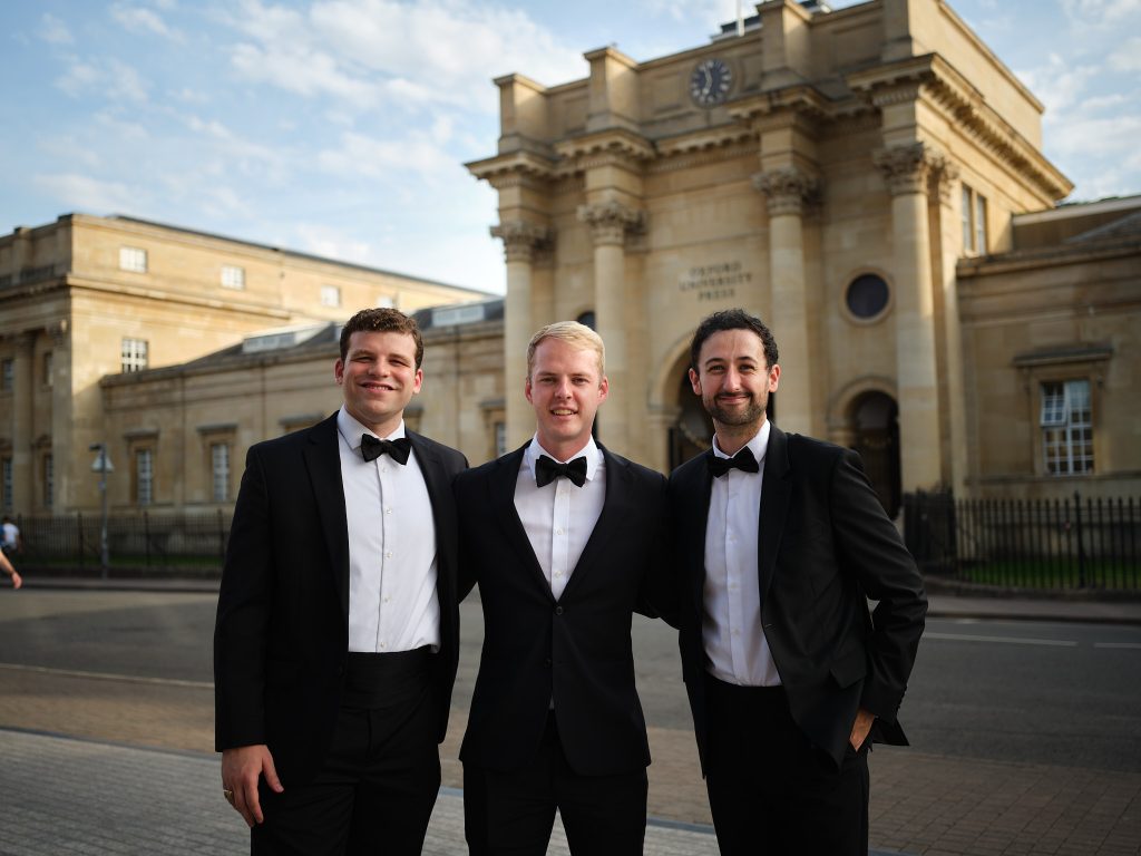 John Richardson and two additional Oxford students stand together in tuxedos with black bowties in from of a historic building.
